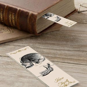Può includere: Un segnalibro beige con un'illustrazione anatomica in bianco e nero di un cranio e di una mascella. Il segnalibro è inserito in un libro marrone. Il testo "Know Thyself" è scritto sul segnalibro in un carattere decorativo.