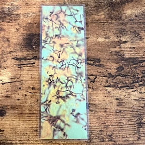 May include: A rectangular bookmark with a floral design. The bookmark features a light blue background with a pattern of yellow and brown branches and flowers. The bookmark is encased in a clear plastic sleeve.