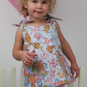 May include: A light blue sleeveless dress with a colourful fish pattern. The dress has tie straps and a pocket with a scale-like design. The dress is worn by a young child.