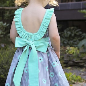 May include: A grey and white striped dress with a mint green bow and trim. The dress has a floral pattern and a ruffled neckline. The back of the dress is visible, showing the bow and button closure. The dress is for a child.