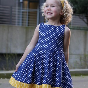 May include: A blue dress with white polka dots and a yellow zigzag hem. The dress is worn by a young person with blonde curly hair and a matching yellow headband. The dress is knee-length.