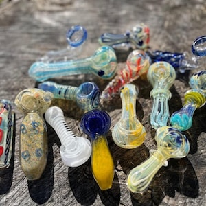 Könnte beinhalten: Eine Sammlung farbenfroher Glaspfeifen in verschiedenen Designs. Die Pfeifen weisen Wirbel aus Blau, Rot, Gelb und Grün auf. Einige haben gemusterte Köpfe und Stiele, während andere klar oder undurchsichtig sind. Die Pfeifen sind auf einer Holzoberfläche angeordnet.