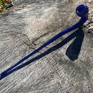 Könnte beinhalten: Eine dunkelblaue Glaspfeife mit gebogenem Stiel und rundem Kopf. Die Pfeife liegt auf einem verwitterten, grauen Baumstumpf. Das Sonnenlicht wirft einen Schatten der Pfeife auf das Holz.