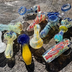May include: A collection of colorful glass pipes in various shapes and designs. The pipes feature swirls of blue, yellow, red, and white, with some having textured or ribbed surfaces. The assortment is displayed on a weathered wooden surface.