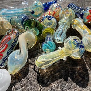 May include: A collection of colorful glass pipes in various shapes and designs. The pipes feature swirls of blue, green, yellow, and red. Some have decorative patterns, while others have a smooth, clear finish. The pipes are arranged on a wooden surface.