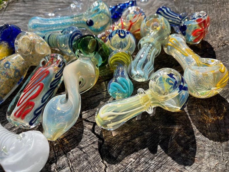 May include: A collection of colorful glass pipes in various shapes and designs. The pipes feature swirls of blue, green, yellow, and red. Some have decorative patterns, while others have a smooth, clear finish. The pipes are arranged on a wooden surface.