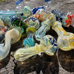 May include: A collection of colorful glass pipes in various designs. The pipes feature swirls of blue, yellow, green, and red, with some having decorative patterns. The glass pipes are displayed on a weathered wooden surface.