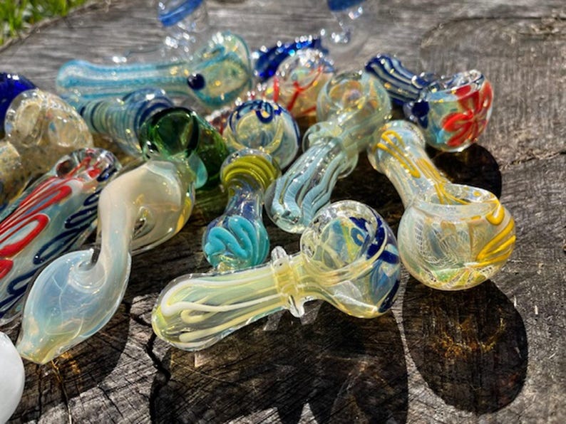 May include: A collection of colorful glass pipes in various designs. The pipes feature swirls of blue, yellow, green, and red, with some having decorative patterns. The glass pipes are displayed on a weathered wooden surface.