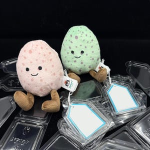 May include: Two plush egg-shaped toys, one pink and one green, each with a smiling face and brown legs. The pink egg has pink spots, and the green egg has purple spots. They are sitting on clear plastic packaging.
