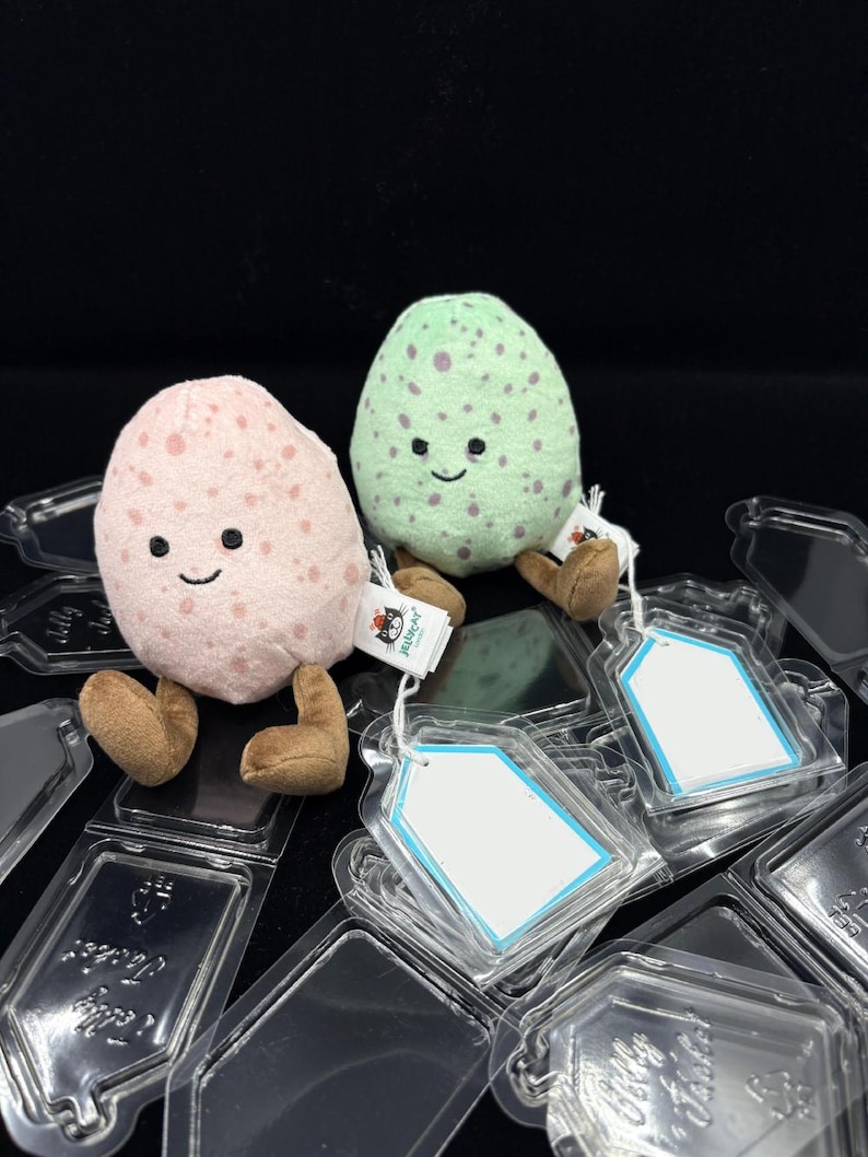 May include: Two plush egg-shaped toys, one pink and one green, each with a smiling face and brown legs. The pink egg has pink spots, and the green egg has purple spots. They are sitting on clear plastic packaging.