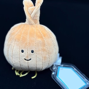 May include: A plush, tan onion with a smiling face and yellow roots. The onion has a textured surface and a twisted top. A clear tag with a blue border is attached to the onion.