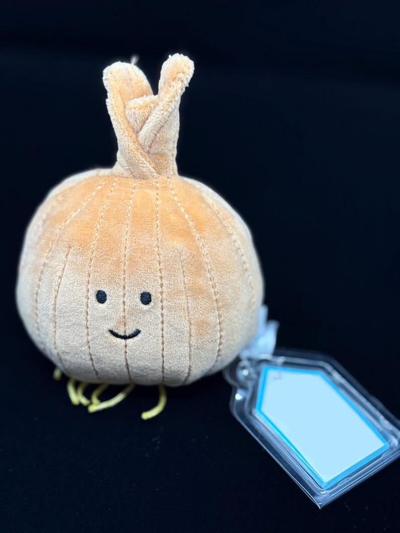 May include: A plush, tan onion with a smiling face and yellow roots. The onion has a textured surface and a twisted top. A clear tag with a blue border is attached to the onion.