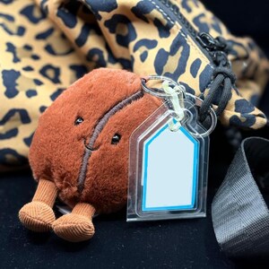May include: A brown plush coffee bean keychain with a smiling face and small legs. The keychain has a clear tag with a blue border. A leopard print bag is in the background.