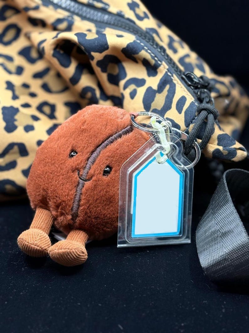 May include: A brown plush coffee bean keychain with a smiling face and small legs. The keychain has a clear tag with a blue border. A leopard print bag is in the background.