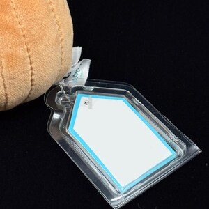 May include: A plush, tan-colored stuffed animal with a clear plastic tag attached. The tag has a white insert with a light blue border. The background is black.