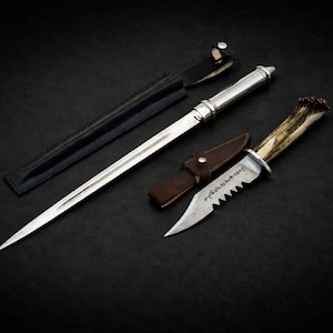 May include: An assortment of handcrafted knives and a sword, displayed on a dark surface. The sword has a silver handle and a black sheath. One knife features a serrated blade and a handle made of antler. A brown leather sheath is included.