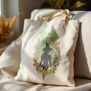 Puede incluir: Un bolso tote color crema con una silueta enmarcada de una bruja con sombrero y vestido. El marco est&aacute; decorado con flores y un lazo verde. El bolso tiene dos asas y est&aacute; hecho de un material similar a la lona.