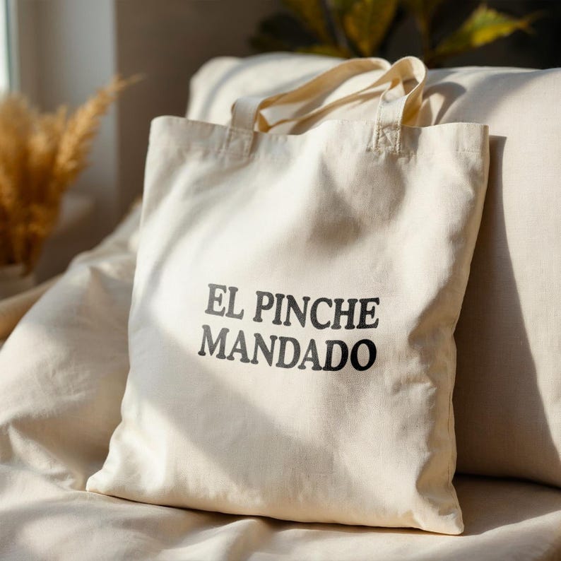 May include: Cream-colored tote bag with the text "EL PINCHE MANDADO" printed in black. The bag has two handles and appears to be made of canvas or a similar fabric. The bag is sitting on a light-colored surface.