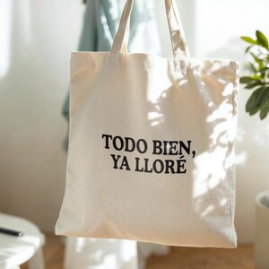 May include: Cream-colored tote bag with the text "TODO BIEN, YA LLOR&Eacute;" in black. The bag has two handles and is hanging in front of a light-colored wall. The bag is made of a sturdy fabric, suitable for carrying groceries or personal items.