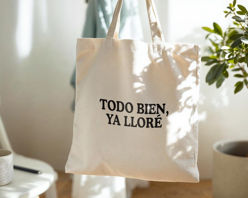 May include: Cream-colored tote bag with the text "TODO BIEN, YA LLOR&Eacute;" in black. The bag has two handles and is hanging in front of a light-colored wall. The bag is made of a sturdy fabric, suitable for carrying groceries or personal items.