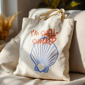 Puede incluir: Bolso tote de color crema con el texto "I'M SHELL-SHOCKED" en rojo y un diseño de concha azul. El bolso tiene dos asas y está hecho de una tela resistente. El diseño es sencillo y llamativo, perfecto para llevar la compra o artículos personales.