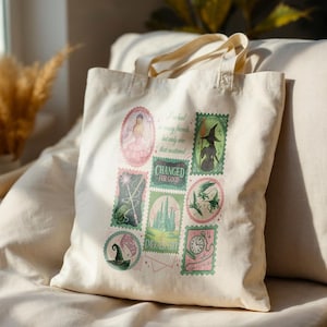 Könnte beinhalten: Cremefarbene Tragetasche mit einem farbenfrohen, stempelartigen Design mit Bildern und Text aus dem Zauberer von Oz. Die Stempel zeigen eine Hexe, ein Schloss und die Worte "Emerald City" und "Changed for Good."