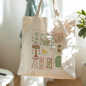 May include: A cream-coloured tote bag with a colourful collage of illustrations. The design includes a lamp, a duck, a teapot, and a jar of cookies, all within decorative frames. The words "Green Era" and "In My" are also visible.