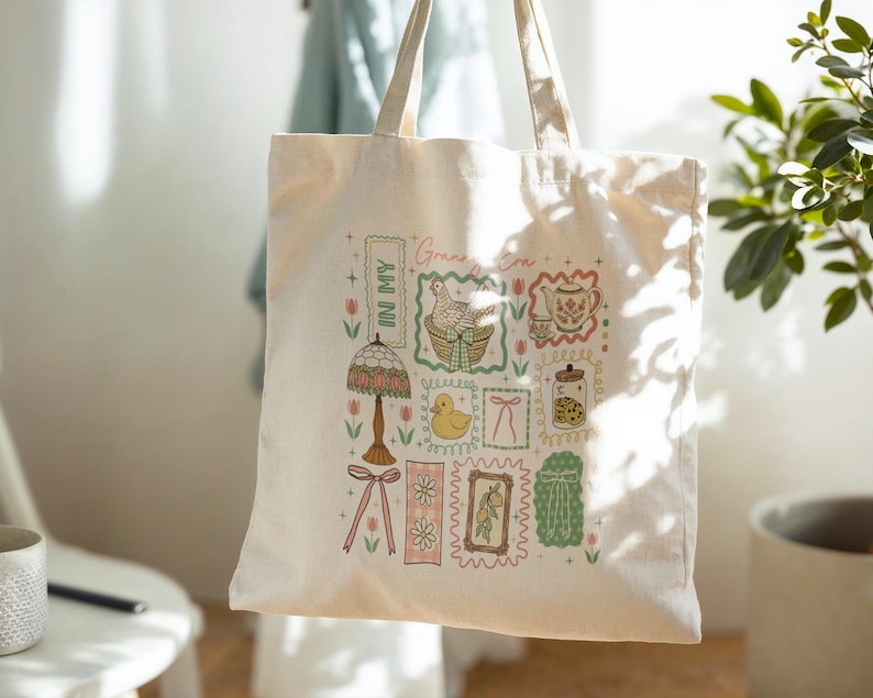 May include: A cream-coloured tote bag with a colourful collage of illustrations. The design includes a lamp, a duck, a teapot, and a jar of cookies, all within decorative frames. The words "Green Era" and "In My" are also visible.