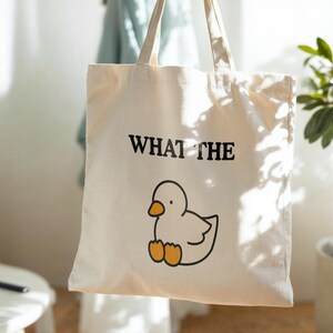 May include: Cream-colored tote bag with black text that reads "WHAT THE" above a cartoon duck illustration. The duck is white with an orange beak and feet. The bag has long, cream-colored handles.