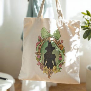 Puede incluir: Bolso tote color crema con una silueta enmarcada de una bruja con sombrero y vestido. El marco est&aacute; rodeado de flores y una cinta verde. El bolso tiene dos asas y cuelga frente a un fondo claro.