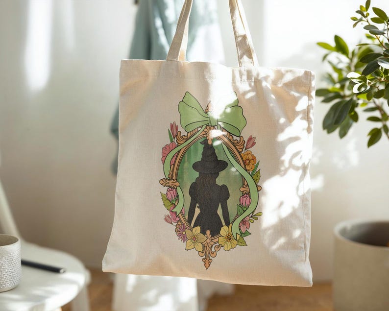 Puede incluir: Bolso tote color crema con una silueta enmarcada de una bruja con sombrero y vestido. El marco est&aacute; rodeado de flores y una cinta verde. El bolso tiene dos asas y cuelga frente a un fondo claro.