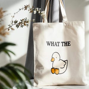 May include: A cream-colored tote bag with the phrase "WHAT THE" above a cartoon duck illustration. The duck is white with an orange beak and feet. The bag has a simple design and is made of a canvas-like material.