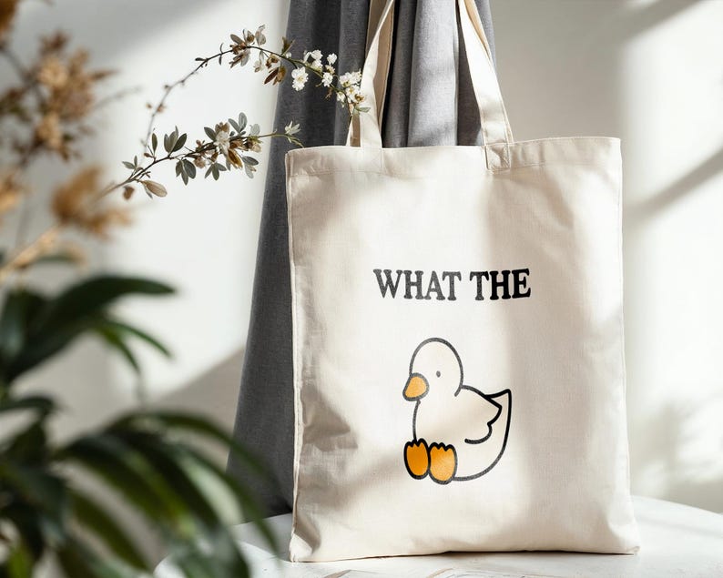 May include: A cream-colored tote bag with the phrase "WHAT THE" above a cartoon duck illustration. The duck is white with an orange beak and feet. The bag has a simple design and is made of a canvas-like material.