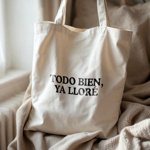 May include: Off-white canvas tote bag with black text that reads "TODO BIEN, YA LLOR&Eacute;". The bag has two handles and is sitting on a beige blanket. The background includes a window and a vase.