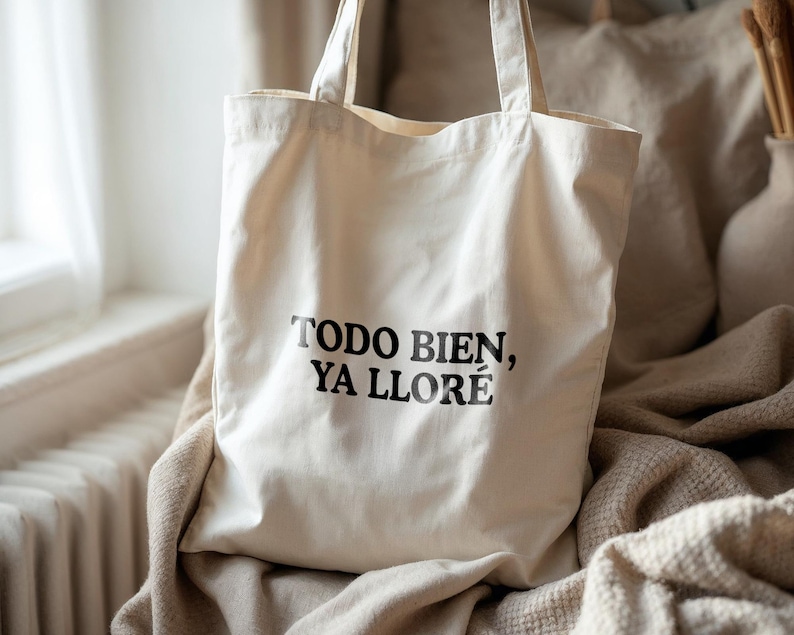 May include: Off-white canvas tote bag with black text that reads "TODO BIEN, YA LLOR&Eacute;". The bag has two handles and is sitting on a beige blanket. The background includes a window and a vase.