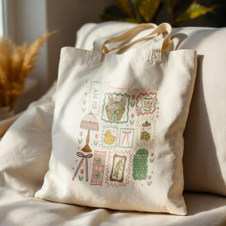 May include: Cream tote bag with a colourful graphic design. The design includes the text "Granny Era" and "In My", plus illustrations of a lamp, duck, teapot, cookies, and other vintage-style elements. The bag has a simple, practical design.