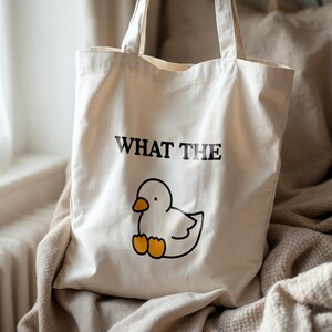 May include: Cream-colored tote bag with the phrase "WHAT THE" above a cartoon duck illustration. The duck is white with a black outline and orange feet. The bag has two handles and is made of a canvas-like material.
