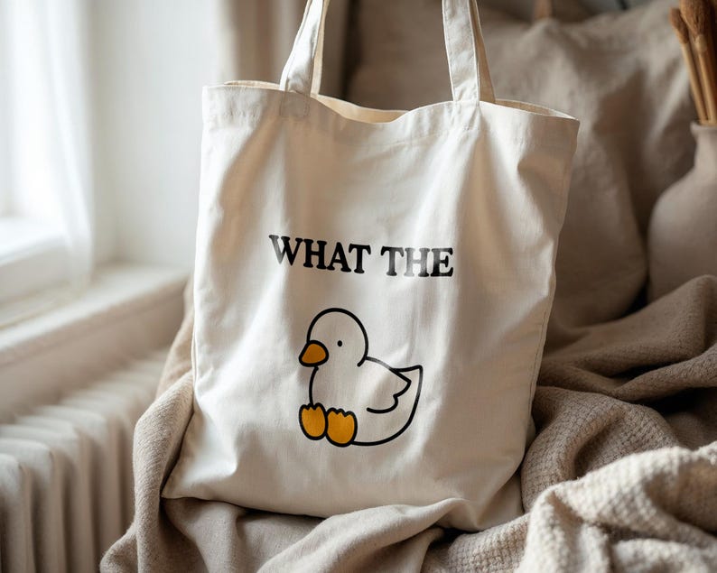 May include: Cream-colored tote bag with the phrase "WHAT THE" above a cartoon duck illustration. The duck is white with a black outline and orange feet. The bag has two handles and is made of a canvas-like material.