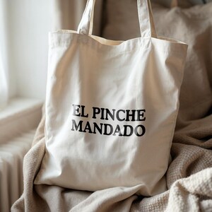 May include: Cream-colored canvas tote bag with the text "EL PINCHE MANDADO" printed in black. The bag has two handles and is sitting on a beige blanket. The background includes a window and a vase.