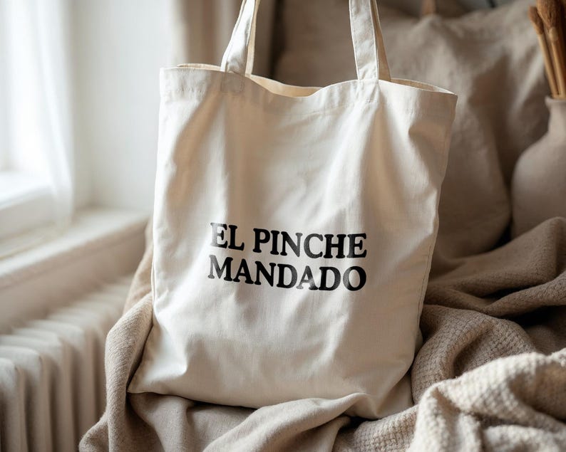 May include: Cream-colored canvas tote bag with the text "EL PINCHE MANDADO" printed in black. The bag has two handles and is sitting on a beige blanket. The background includes a window and a vase.