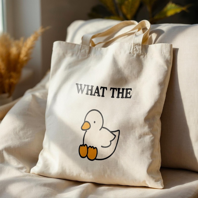 May include: Cream-colored tote bag with the phrase "WHAT THE" printed in black above a cartoon duck illustration. The duck has a white body, orange beak, and orange feet. The bag has two handles and is made of a soft fabric.
