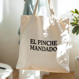 May include: Cream-colored tote bag with the text "EL PINCHE MANDADO" in black. The bag has two handles and is made of a sturdy fabric. The bag is hanging in front of a light-colored wall.