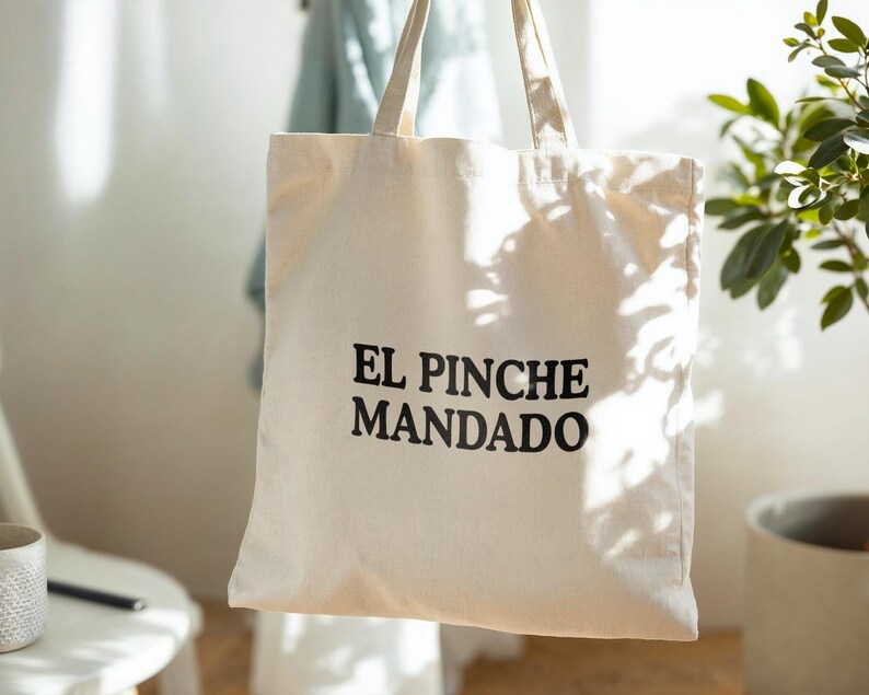 May include: Cream-colored tote bag with the text "EL PINCHE MANDADO" in black. The bag has two handles and is made of a sturdy fabric. The bag is hanging in front of a light-colored wall.