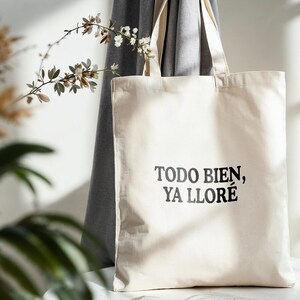 May include: A cream-colored tote bag with the text "TODO BIEN, YA LLOR&Eacute;" in black. The bag is made of a sturdy fabric and has two handles. The background includes a gray curtain and decorative plants.