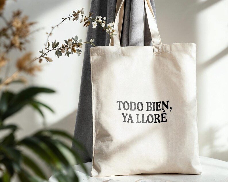 May include: A cream-colored tote bag with the text "TODO BIEN, YA LLOR&Eacute;" in black. The bag is made of a sturdy fabric and has two handles. The background includes a gray curtain and decorative plants.