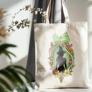 Puede incluir: Un bolso tote de color crema con la silueta de una mujer con sombrero y vestido, enmarcada por un lazo verde, flores y un borde dorado adornado. El bolso tiene asas largas y est&aacute; hecho de una tela resistente.