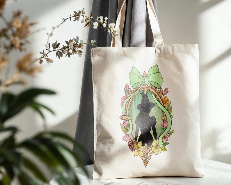 Puede incluir: Un bolso tote de color crema con la silueta de una mujer con sombrero y vestido, enmarcada por un lazo verde, flores y un borde dorado adornado. El bolso tiene asas largas y est&aacute; hecho de una tela resistente.