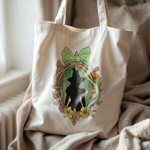 Puede incluir: Bolso tote de lona color crema con una silueta enmarcada de una bruja con sombrero y vestido. El marco est&aacute; decorado con un lazo verde y flores coloridas. El bolso tiene dos asas para llevar.