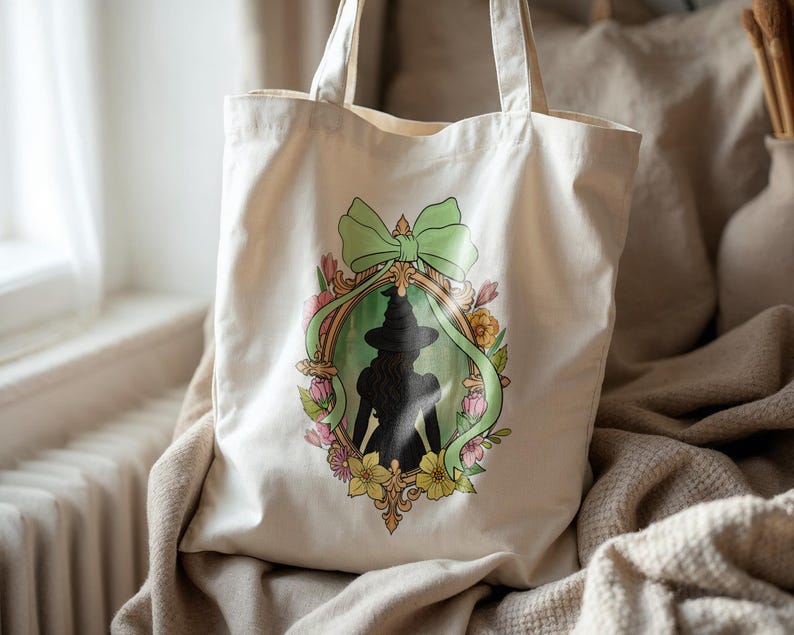 Puede incluir: Bolso tote de lona color crema con una silueta enmarcada de una bruja con sombrero y vestido. El marco est&aacute; decorado con un lazo verde y flores coloridas. El bolso tiene dos asas para llevar.