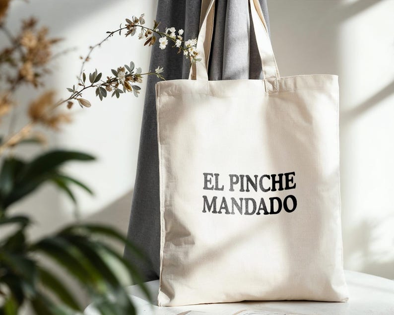 May include: A cream-colored tote bag with the words "EL PINCHE MANDADO" printed in black. The bag has a rectangular shape with two handles and is made of a canvas-like material. The bag is displayed against a neutral background.